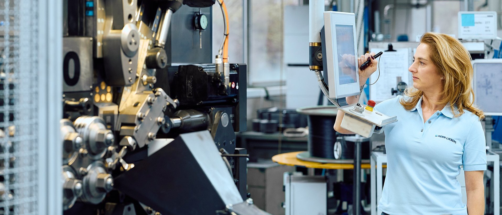 Frau steht an einem Windeautomat, welcher eine Druckfeder herstellt und prüft die Qualität der Drahtfeder.